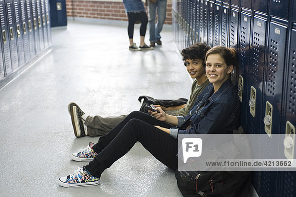 Gymnasiast sitzend auf dem Boden mit Freund bei Schließfächern im Schulflur