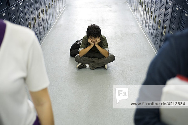 Teenage boy sitting sullenly on floor of school corridor  classmates in foreground