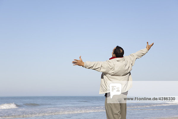 Mann steht am Strand und schaut mit ausgestreckten Armen auf den Ozean.