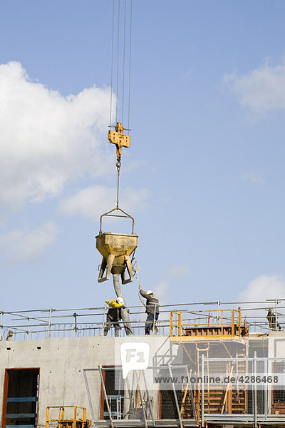 Bauarbeiter  die mit einem Kran hängende Schaufel führen  um Beton auf ein im Bau befindliches Gebäude zu legen.
