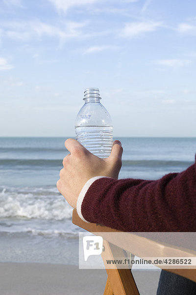 Person sitzt entspannt am Strand  Wasserflasche in der Hand