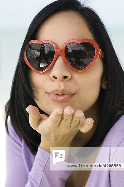 Frau mit herzförmiger Sonnenbrille  die einen Kuss vor die Kamera bläst.