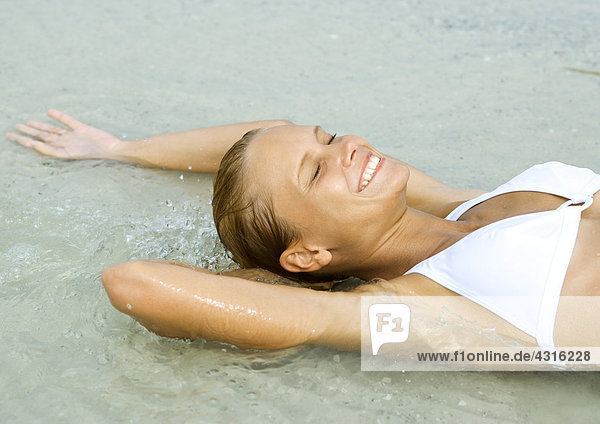 Frau liegt im flachen Wasser am Strand und lächelt mit geschlossenen Augen.
