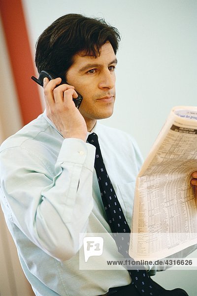 Businessman using phone and holding up financial section of newspaper