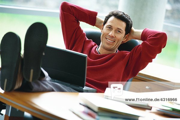 Mann sitzt am Schreibtisch mit erhobenen Füßen  hält Laptop auf dem Schoß  Hände hinter dem Kopf