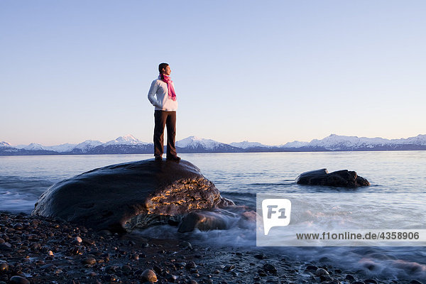 Woman stands on rock at Bishops Beach viewing Kachemak Bay in Homer  Kenai Peninsula  Southcentral Alaska  Winter