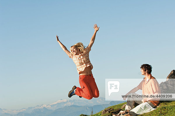 Mann sitzend  Frau springt in die Luft  Arme hoch  lächelnd.
