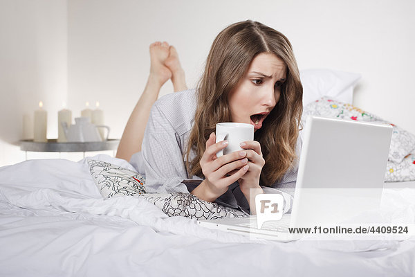 Woman lying on bed holding coffee cup with laptop  mouth open