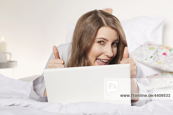Woman lying on bed with laptop and showing thumbs up