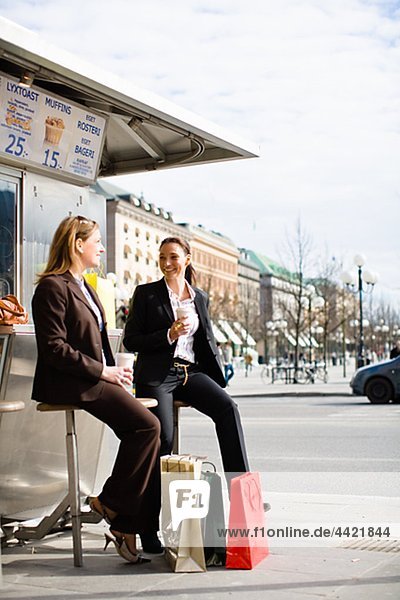 Paar von eleganten Frauen warten auf Bushaltestelle mit Einkaufstaschen