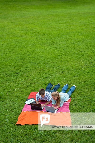 Mid adult couple lying on lawn in park and using laptops