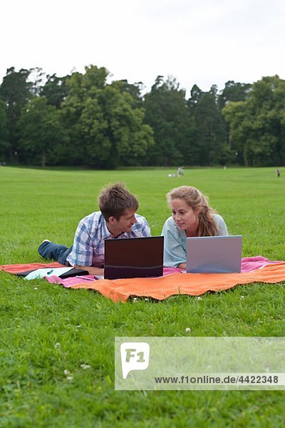 Mid adult couple lying on blanket in park and using laptops