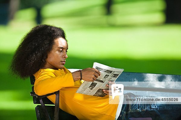 Junger Mann mit Afro Haar lesen Zeitung auf Bank