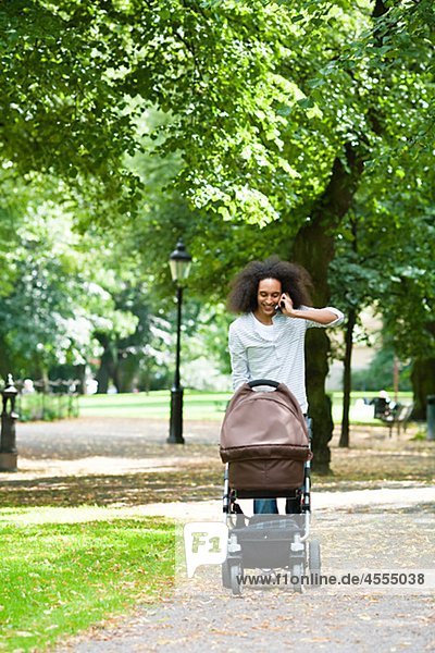 Young man pushing baby carriage in park and talking on mobile phone