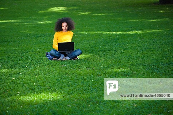 Young man with afro hair sitting on grass with laptop