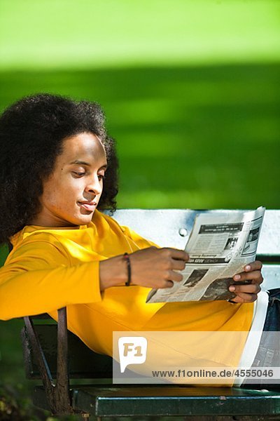 Young man with afro hair reading newspaper on bench