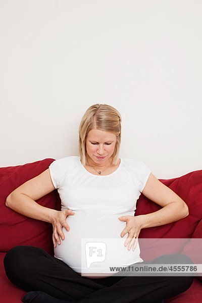 Schwangere Frau sitzt auf Sofa mit Händen auf Bauch