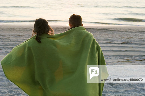 Children wrapped together in blanket standing on beach observing sunset