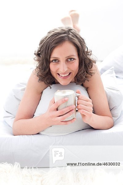 Junge Frau auf dem Bett liegend mit Kaffeetasse  lächelnd  Porträt