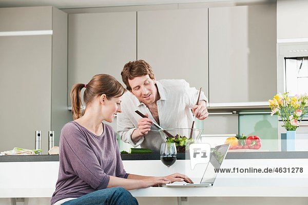 Mann bereitet Salat mit Frau mit Laptop in der Küche zu