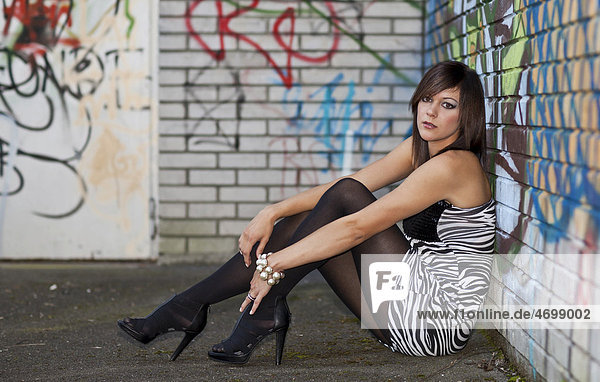 Young woman with dark hair wearing a top with a zebra pattern and high heels posing while sitting in front of a wall with graffiti