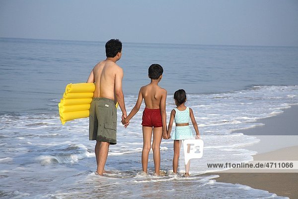 South Asian Indian father and children holding hands carrying yellow floating air bed in arm standing in surf water on seashore   Shiroda   Dist Sindhudurga   Maharashtra   India MR703D 703F 703G