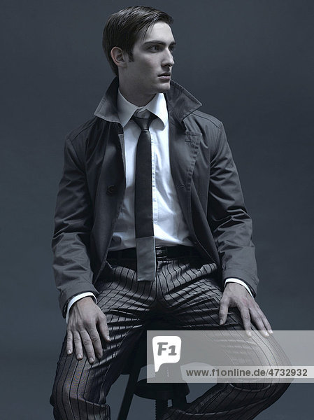 Modeaufnahme eines Mannes im Anzug und Mantel  auf Hocker sitzend