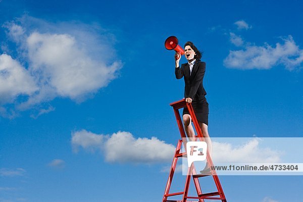 Businesswoman standing on top of red ladder against blue sky and shouting through red megaphone