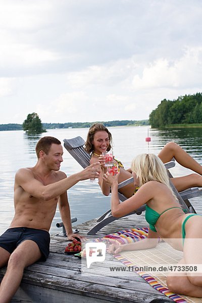 One man and two women sitting on jetty and drinking cocktails