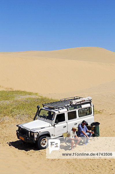 Two men sitting in front of a Landrover Defender off-road vehicle in the dunes of the Namib Naukluft National Park  part of the Namibian Skeleton Coast National Park  Skeleton Coast  Namib Desert  Namibia  Africa