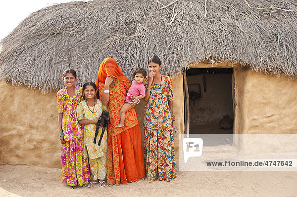 Mädchen und junge Frauen stehen neugierig vor ihrem Wohnhaus  eine junge Frau im roten Sari  verschleiert  trägt ihr Kind auf dem Arm  ein Mädchen hält eine junge Ziege  Wüste Thar  Rajasthan  Indien  Asien