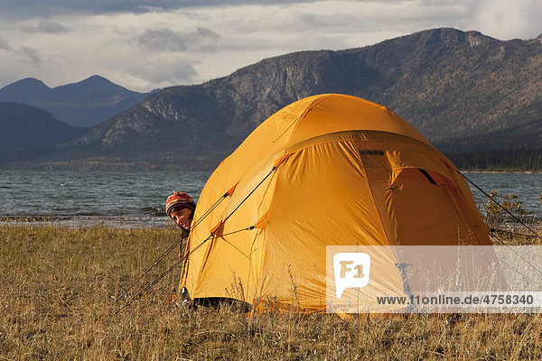Junge Frau schaut aus einem Zelt heraus  am See Kusawa Lake  dahinter die Berge  Yukon Territorium  Kanada