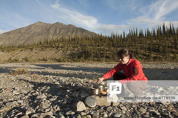 Young woman cooking on a camp fire  camping  gravel bar  Northern Mackenzie Mountains and Wind River behind  Yukon Territory  Canada