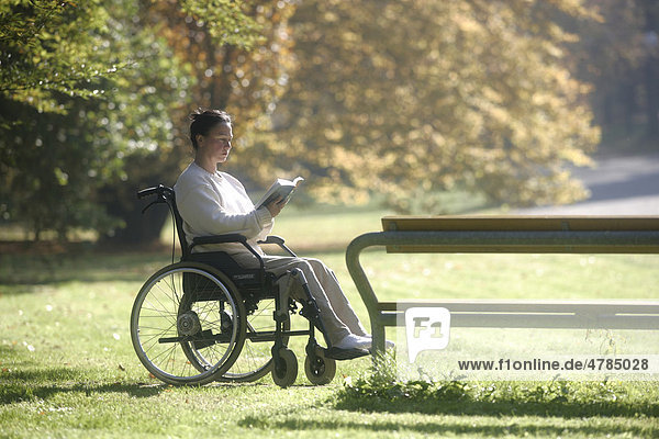Frau liest im Rollstuhl neben Parkbank