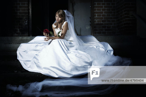Junge Frau in einem Hochzeitskleid und Schleier sitzt in einer urbanen Location auf einer Treppe und hält eine rote Rose in der Hand