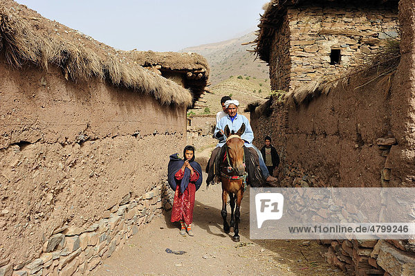 Ein kleines Mädchen mit einem Tragetuch auf dem Rücken und zwei Reiter auf einem Pferd in einer schmalen Gasse eines Dorfes  Wände aus Natursteinen und Stampflehm mit Gras abgedeckt  Kelaa M'gouna  Hoher Atlas  Marokko  Afrika