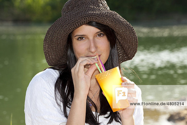 Junge Frau mit Strohhut sitzt am Strand eines Sees und trinkt ein Erfrischungsgetränk