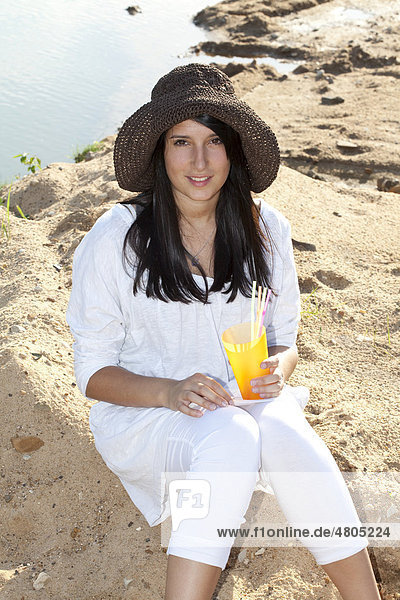 Junge Frau mit Strohhut sitzt am Strand eines Sees und hält ein Erfrischungsgetränk in der Hand