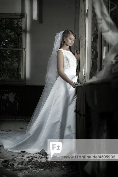 Junge blonde Frau in einem Hochzeitskleid und Schleier sieht in einer urbanen Location aus dem Fenster