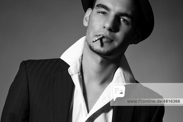 Junger Mann mit Anzug  Hemd  Krawatte und Hut  mit einer Zigarette im Mund und coolem Blick