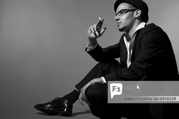 Junger Mann mit Anzug  Hemd  Krawatte und Hut sitzt in Gedanken versunken auf dem Boden und raucht eine Zigarette