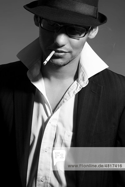 Young man wearing a suit  a shirt  a tie and a hat with a cigarette in his mouth and looking cool