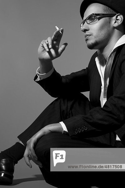 Young man wearing a suit  a shirt and a hat  seated on the ground  lost in thought and smoking a cigarette