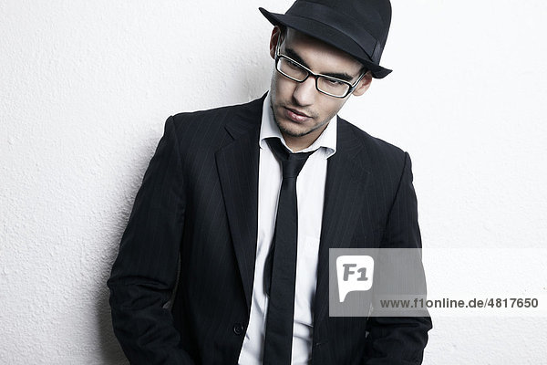 Portrait eines jungen Mannes mit Brille  Anzug  Hemd  Krawatte und Hut angelehnt an eine Wand