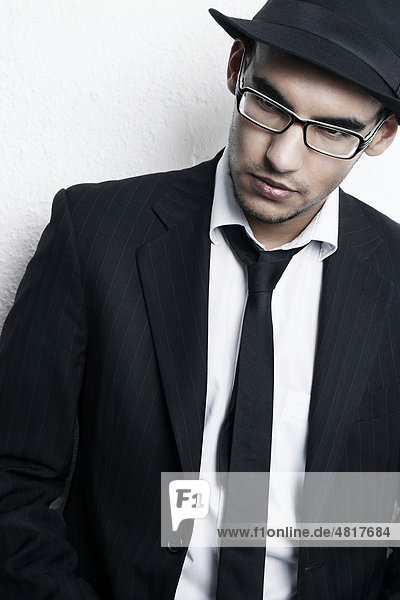 Portrait eines jungen Mannes mit Brille  Anzug  Hemd  Krawatte und Hut angelehnt an eine Wand