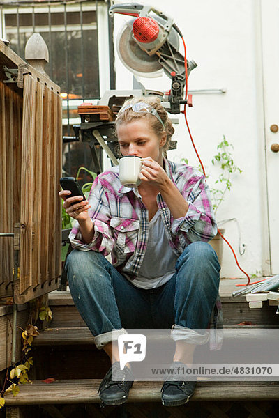 Frau sitzt auf der Treppe mit Smartphone und trinkt heißes Getränk