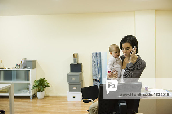 Mutter hält Baby  während sie im Büro telefoniert.