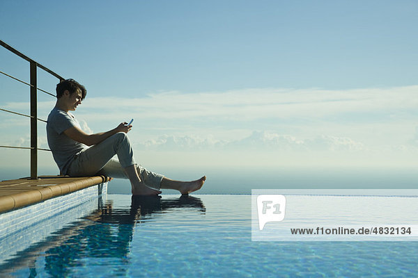 Man sitting on edge of infinity pool  text messaging with cell phone