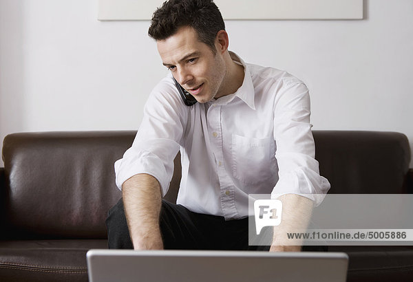Ein Mann  der auf einem schnurlosen Telefon spricht  während er zu Hause einen Laptop benutzt.