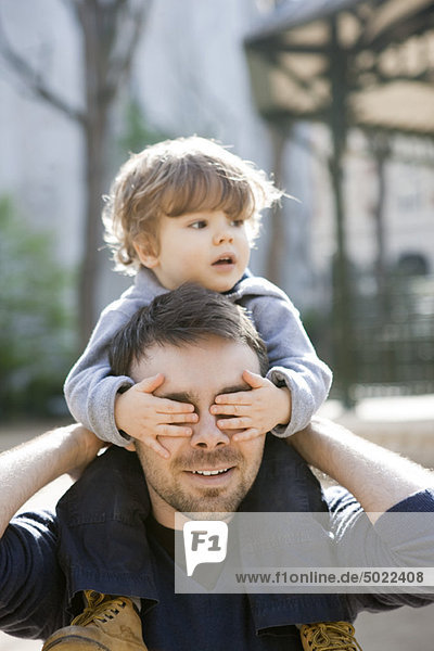 Vater trägt Kleinkindesohn auf seinen Schultern  Sohn bedeckt Vaters Augen mit seinen Händen.